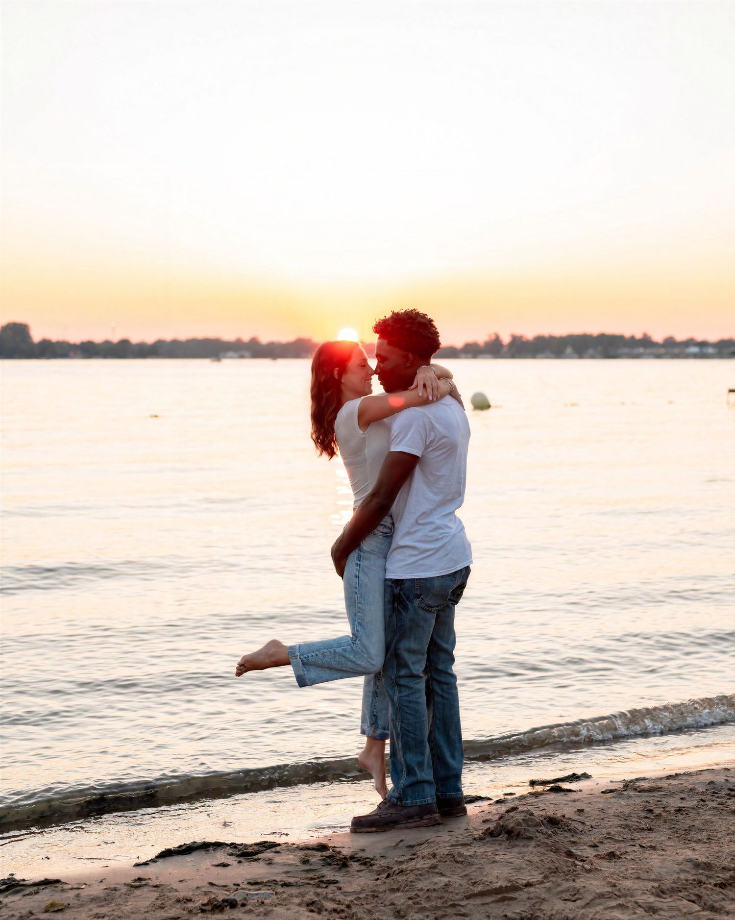 Couple sharing a romantic kiss on a sandy beach at sunset, with calm water and a glowing horizon in the background.