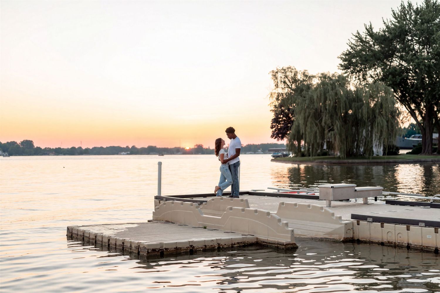 Couple embracing on a dock at sunset over a calm lake with willow trees in the background.
