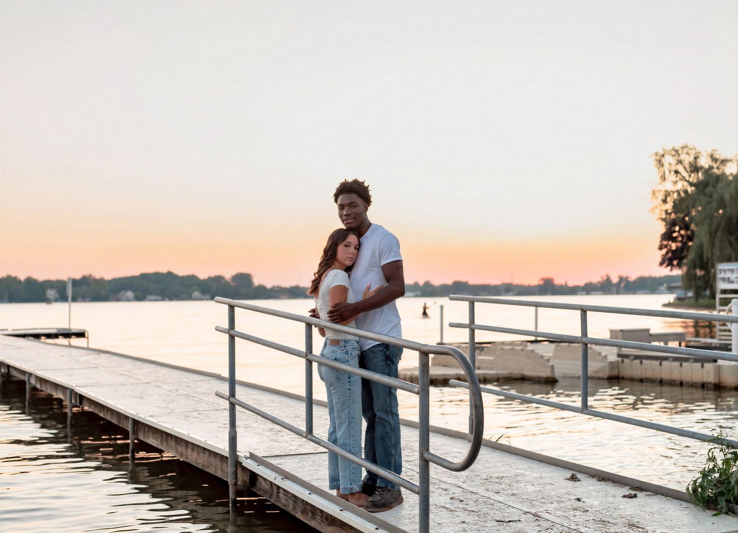 Couple embracing on a lakeside dock at sunset, with calm water and trees in the warm golden background.