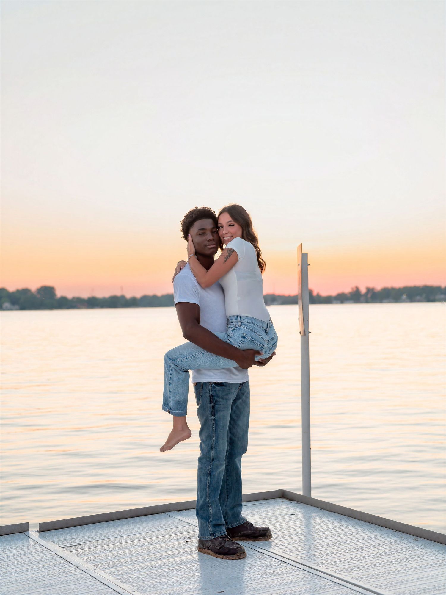 Couple embracing on a dock at sunset, man lifting woman in white tops and jeans with a lake in the background.
