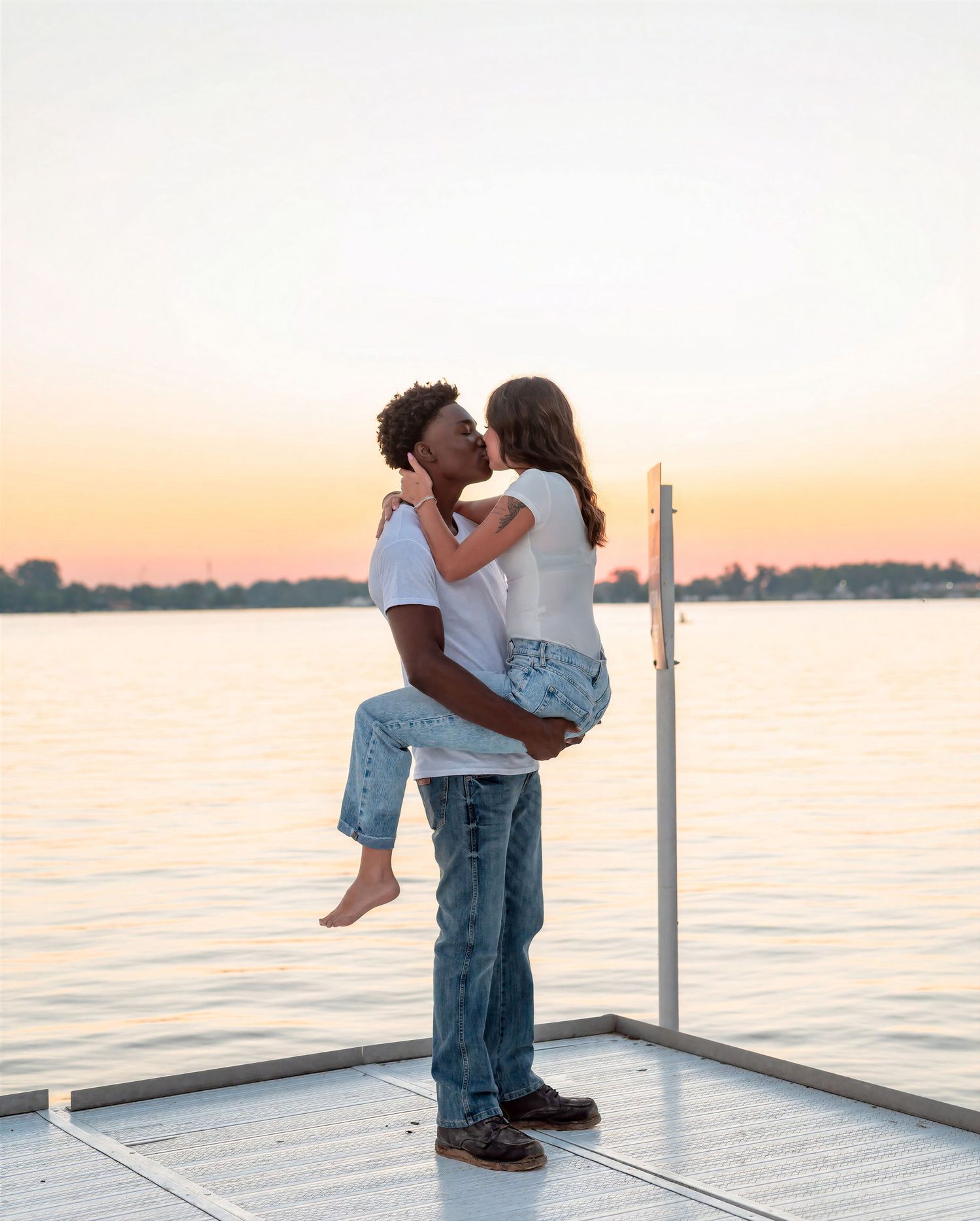 Couple sharing a romantic kiss on a dock at sunset, man lifting woman, with calm water in the background.