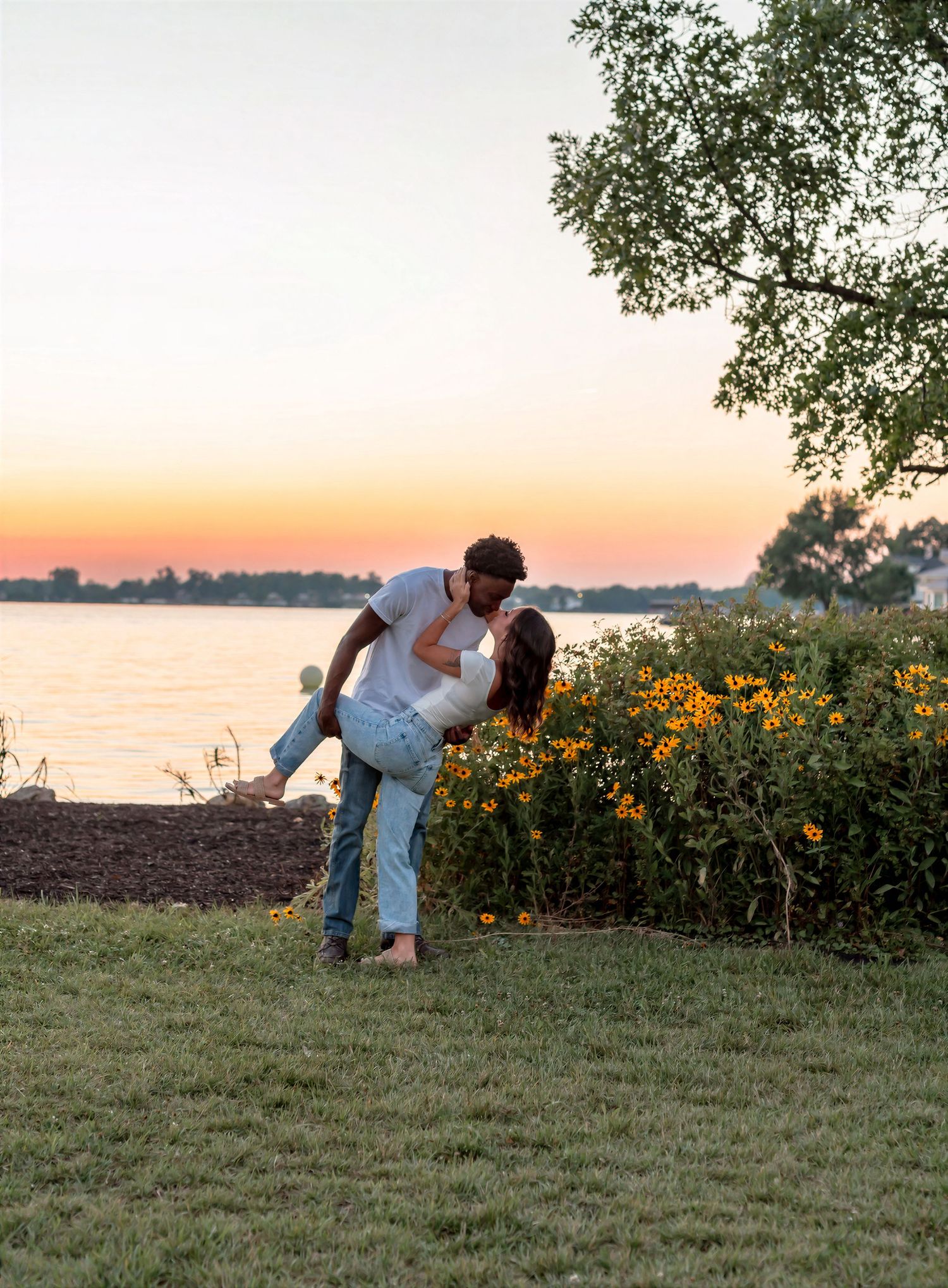 Couple sharing a romantic dip kiss near a lakeside garden with yellow wildflowers during a golden sunset.