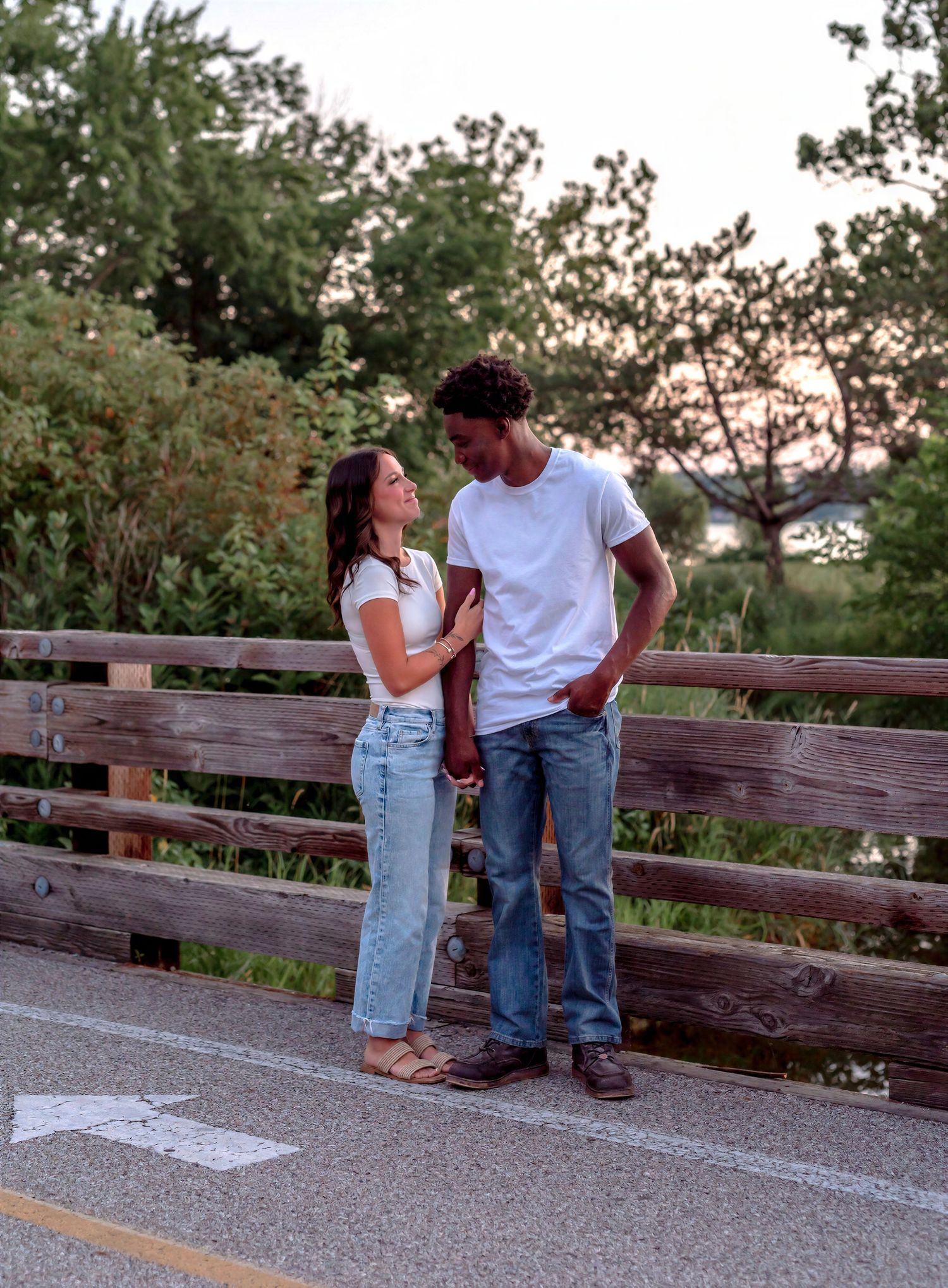 A couple stands on a wooden bridge, smiling at each other, surrounded by lush green trees at sunset.