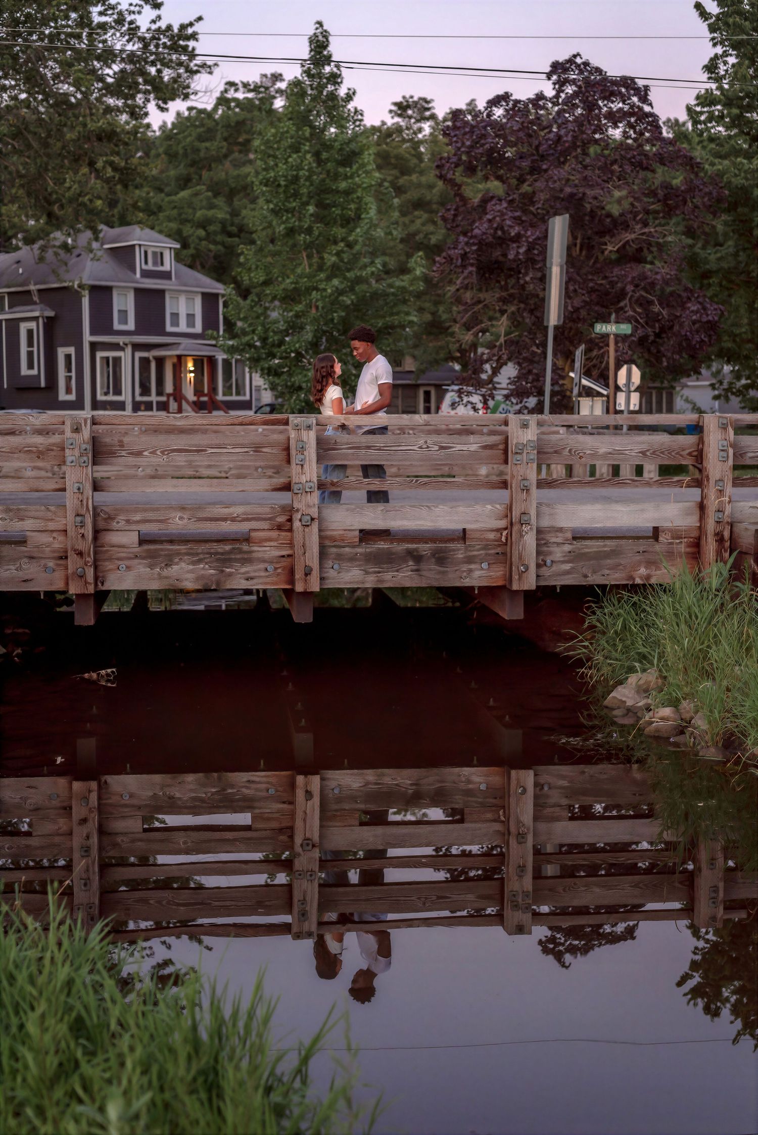 Couple standing on a rustic wooden bridge reflected in calm water at dusk with trees and houses in background.