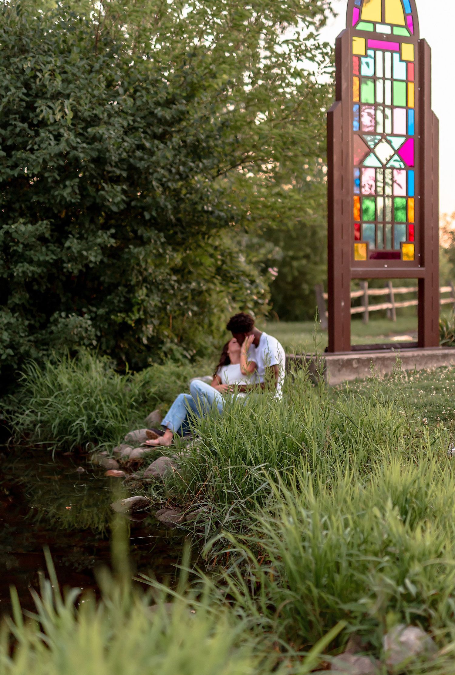 Couple sitting by a creek near a colorful stained glass monument surrounded by lush green trees at sunset.