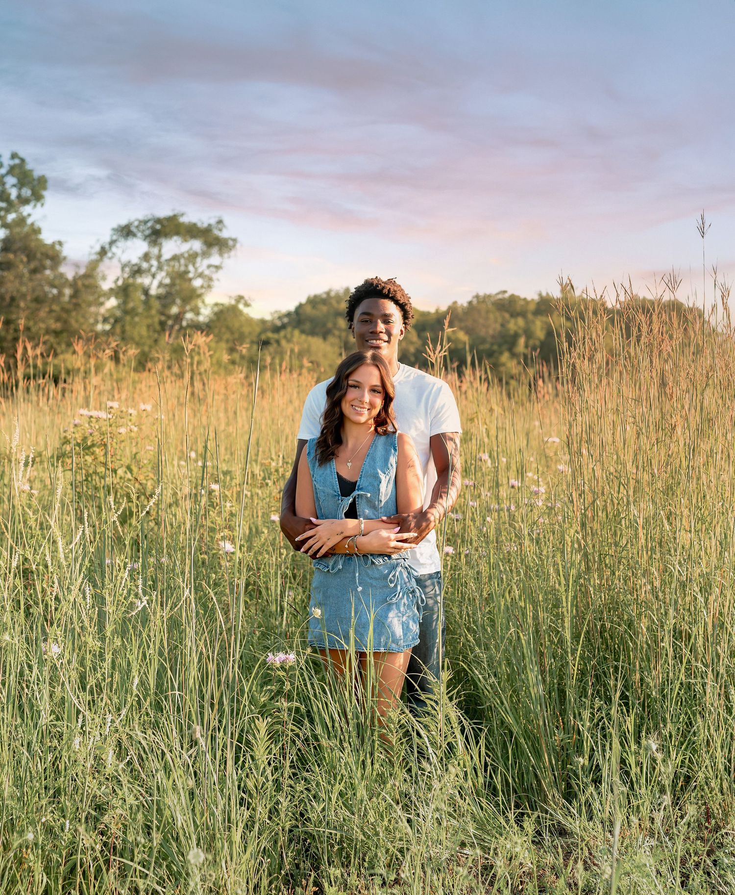 A couple embraces in a lush green meadow at golden hour, surrounded by wildflowers under a pastel sunset sky.