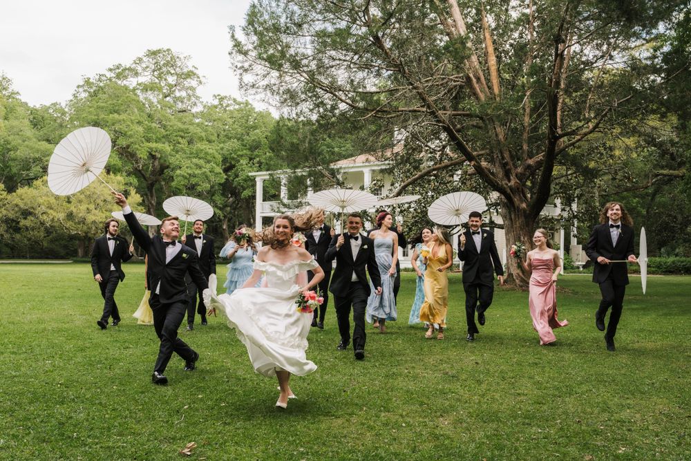 Joyful wedding party running across a green lawn holding white parasols on a cloudy day.