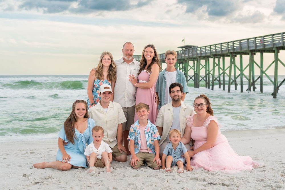 Extended family beach portrait with adults and children posing near a wooden pier at sunset.