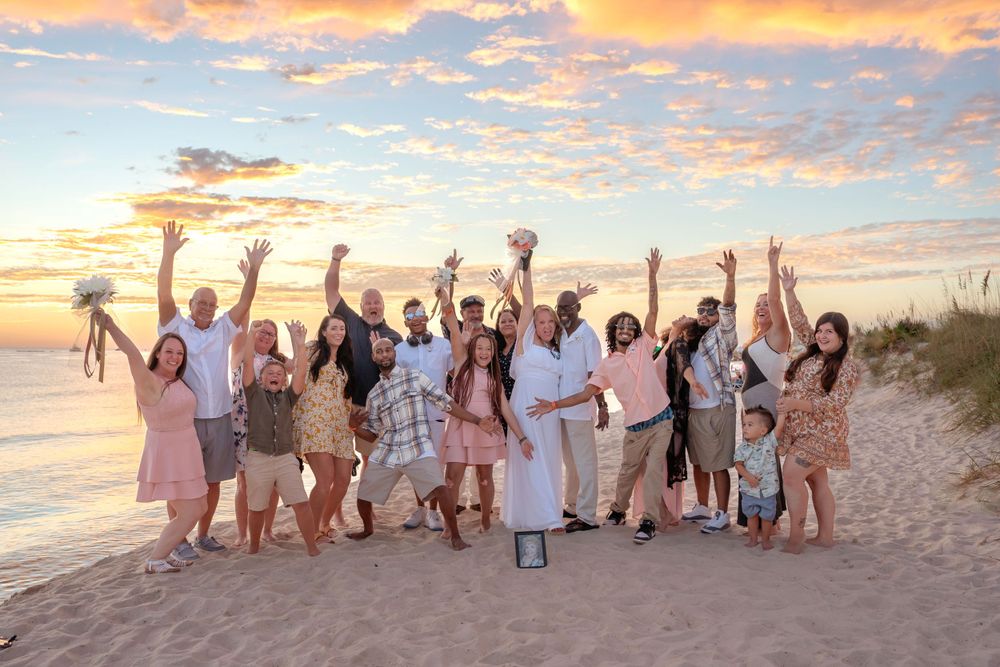 Large wedding party celebrating on a sandy beach at sunset with arms raised in joy.