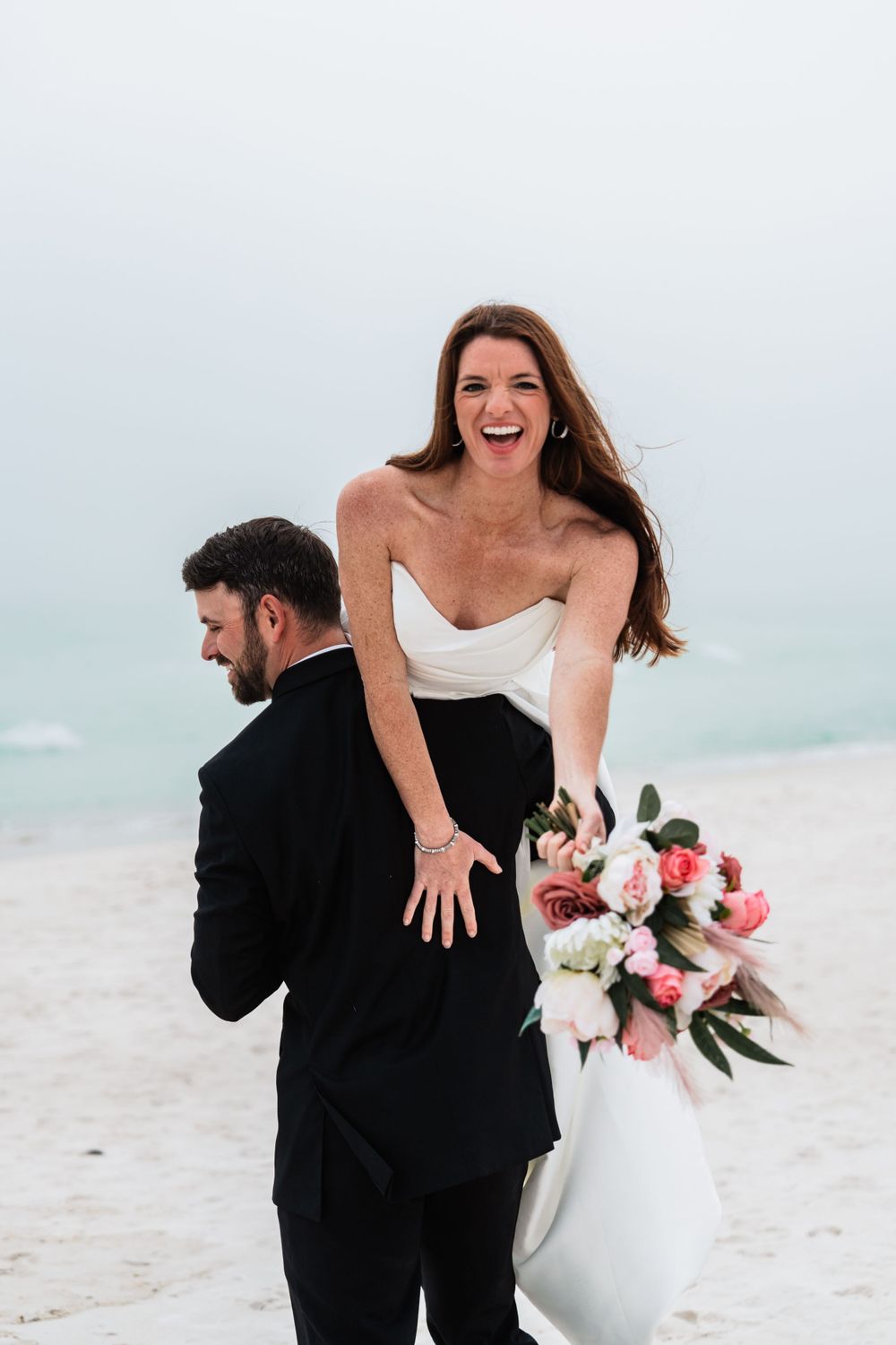 Laughing bride holds a pink bouquet while groom carries her on his back during beach wedding.
