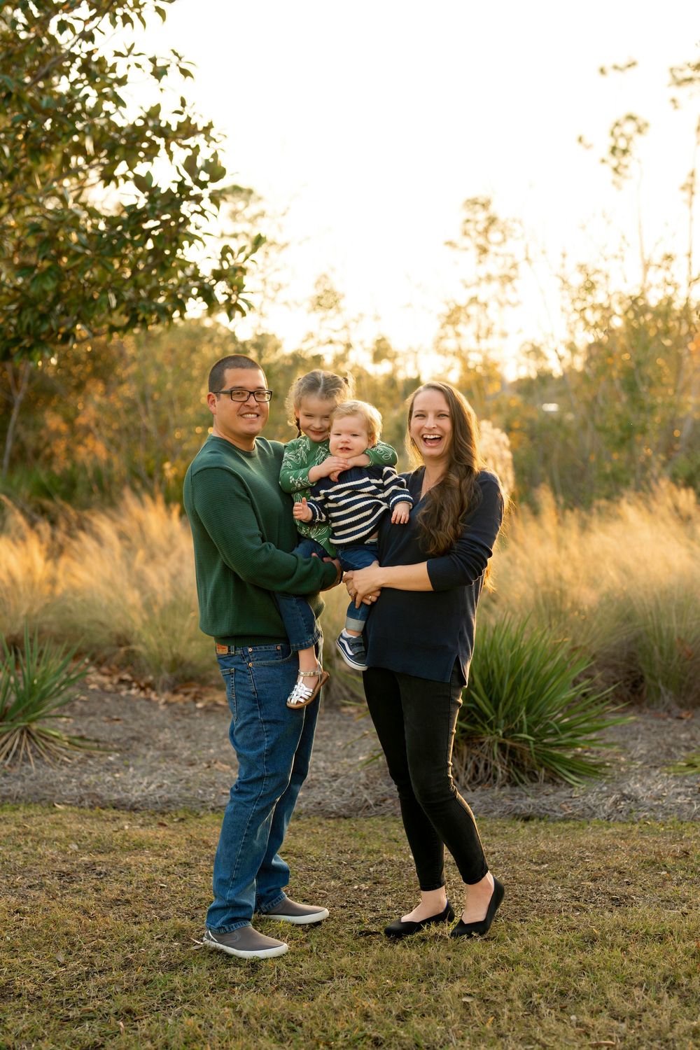 Happy family of four posing outdoors at golden hour, parents holding two young children in a park setting.
