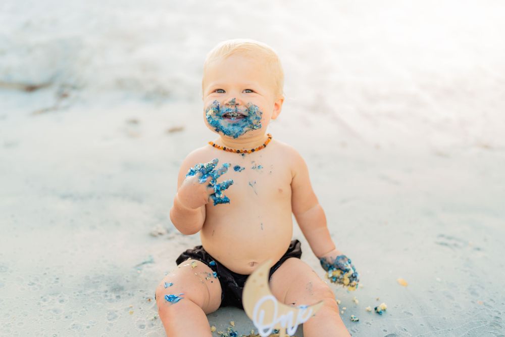 Laughing baby covered in blue frosting sitting on white sand during a beach cake smash session.