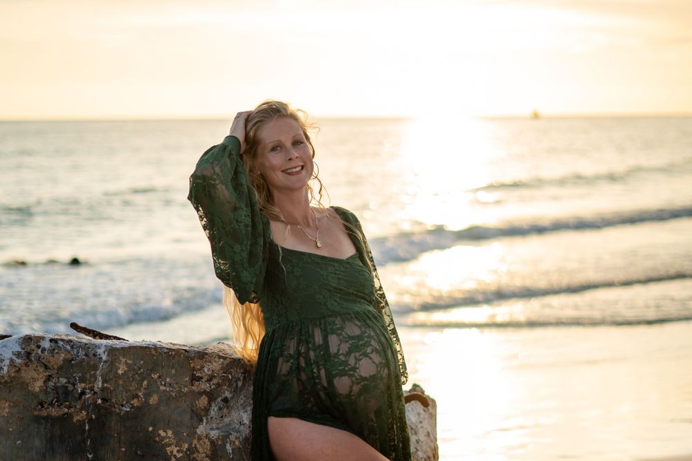 Smiling woman in a green lace cover-up posing on a rock at sunset on the beach.