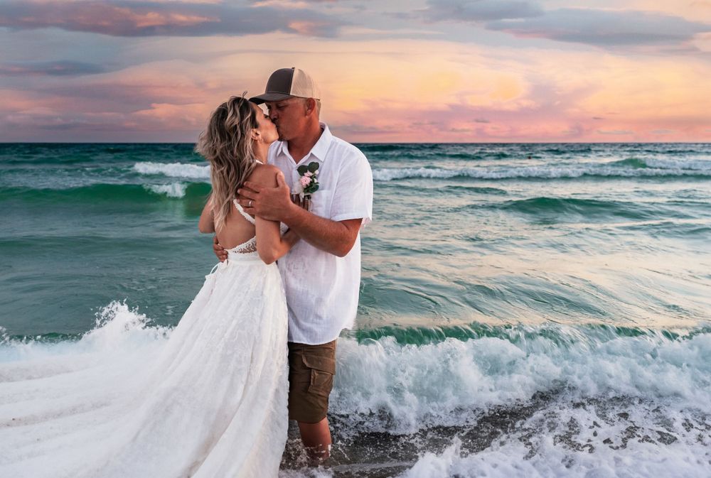 Romantic couple shares a kiss in ocean waves during a stunning colorful beach sunset wedding.