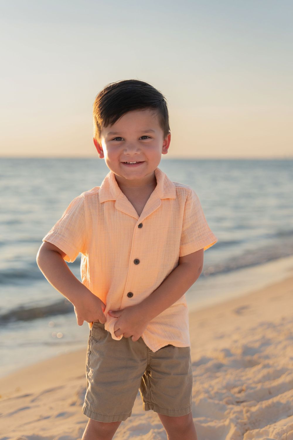 Adorable young boy in a peach shirt smiling on a sandy beach at golden hour.