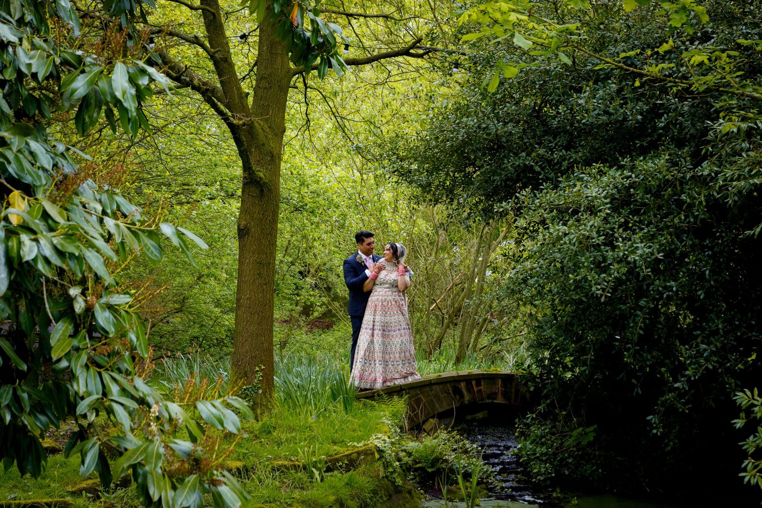 A couple stands on a wooden bridge surrounded by lush spring greenery.