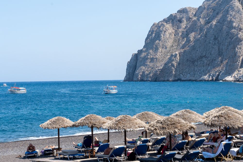 Beach umbrellas and lounge chairs line a Mediterranean coast with towering cliffs and blue waters.