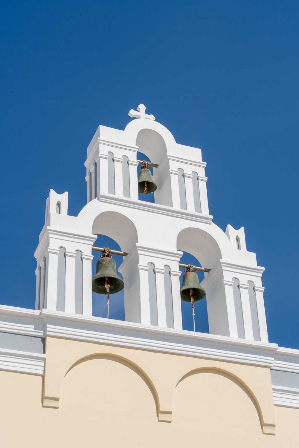 Traditional white Greek bell tower with three bells against vivid blue sky in Santorini.