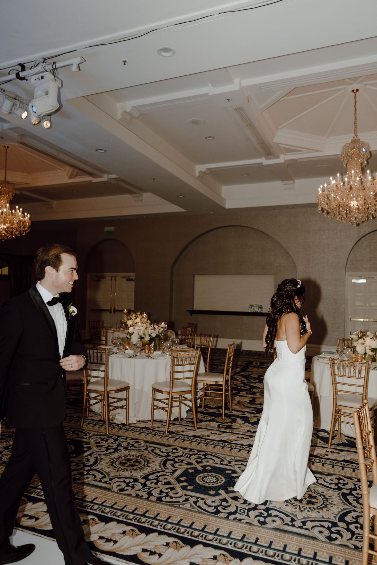 Wedding couple dancing in elegant ballroom with crystal chandeliers and golden chairs.