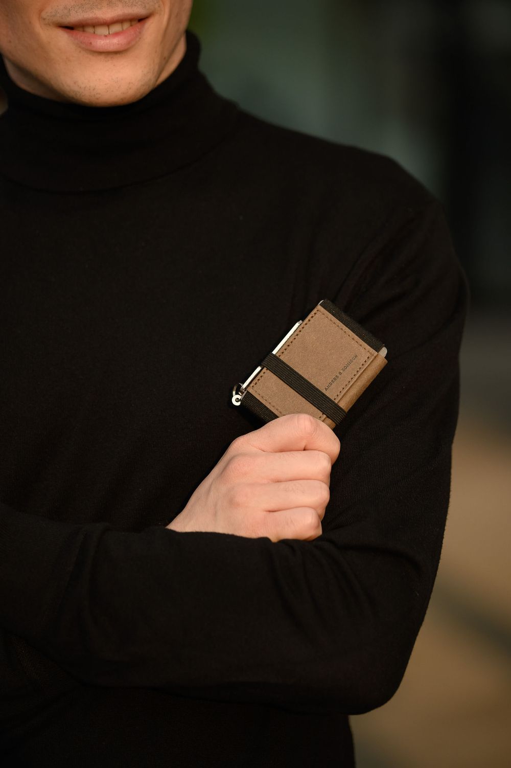 Person in a black turtleneck sweater holds a brown leather wallet against dark background.