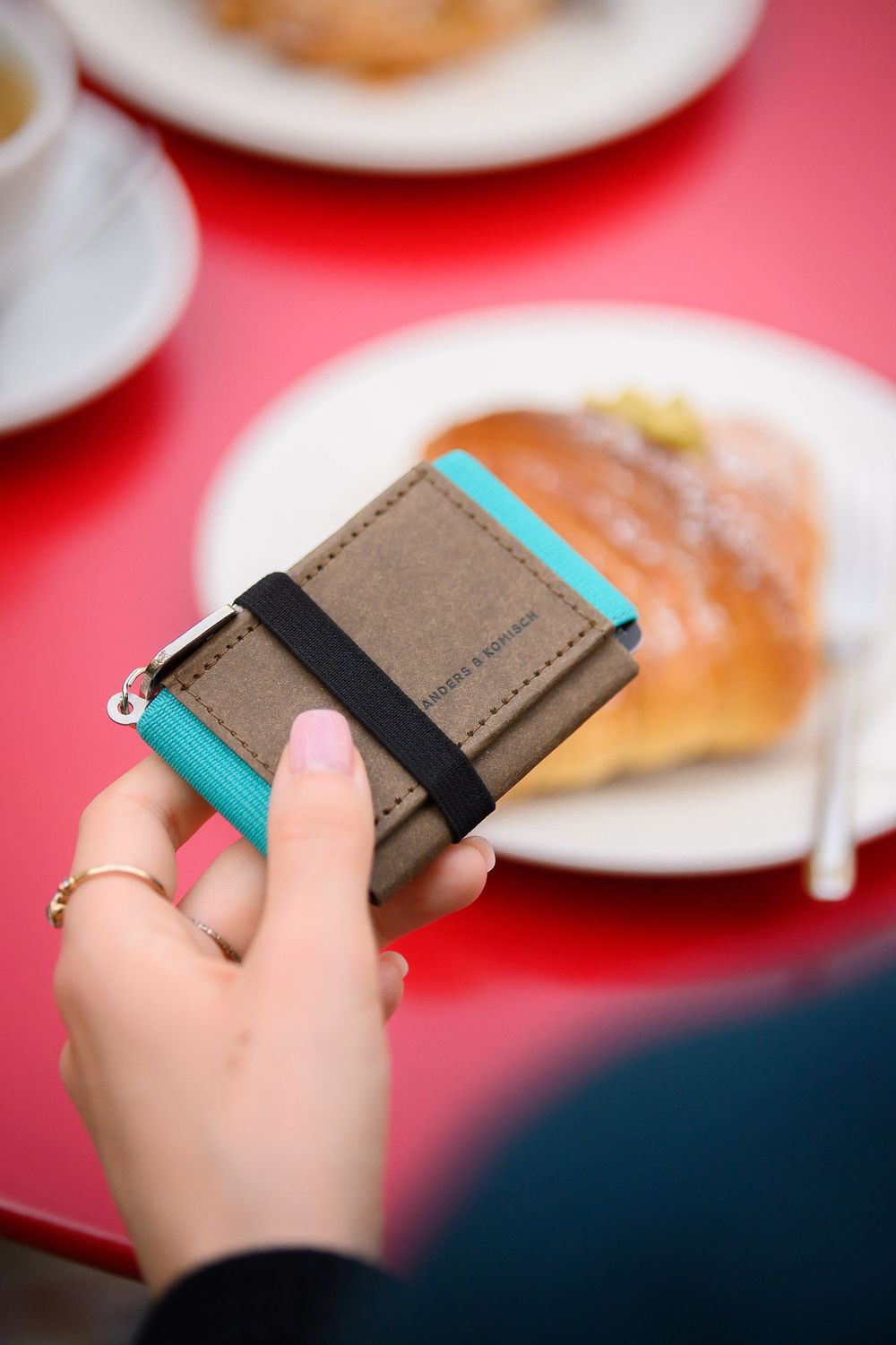 A hand holds a slim brown leather wallet with turquoise accents against a red table background with blurred food plates.