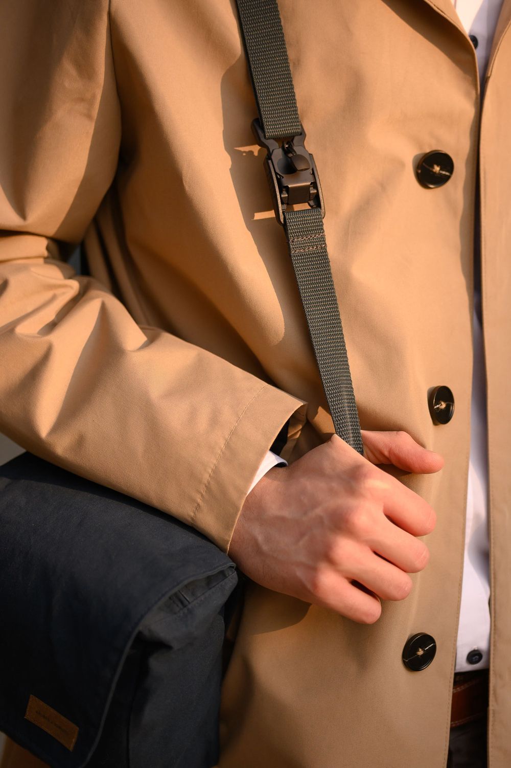 Person in tan trenchcoat adjusting black shoulder strap of messenger bag.