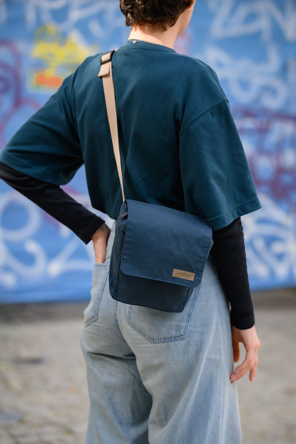 Person wearing navy sweater and jeans with messenger bag against graffiti wall.