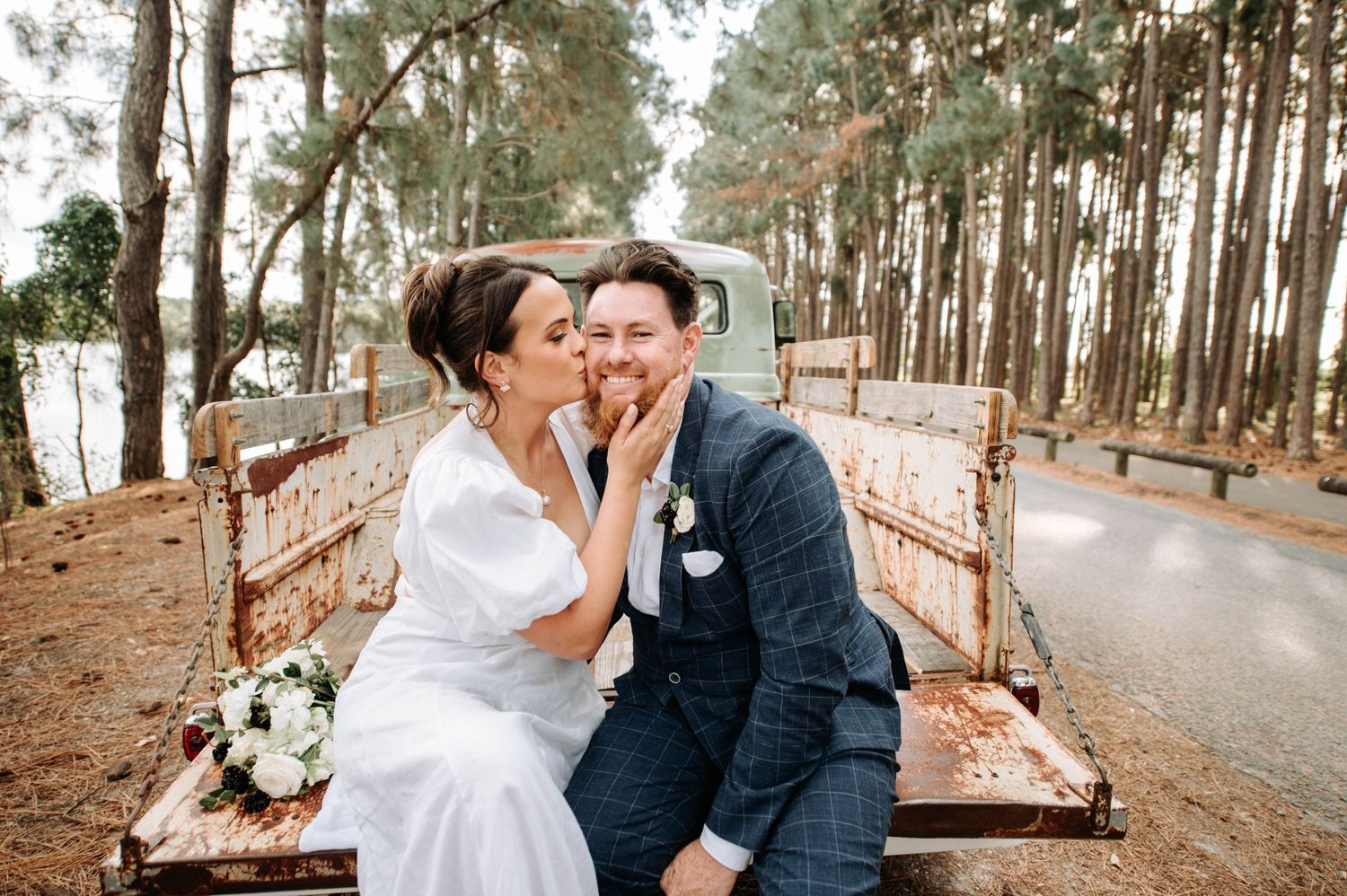 Newlyweds share a joyful moment sitting on vintage truck bed in pine forest setting.