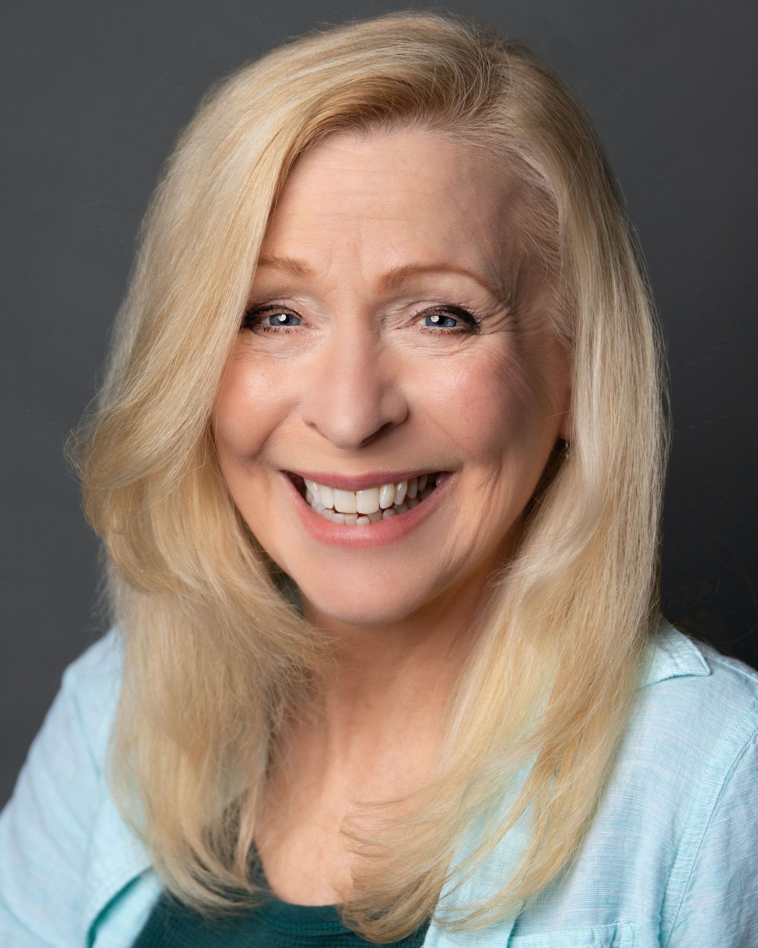 Professional headshot of a blonde woman in a light blue top with a warm smile against dark background.
