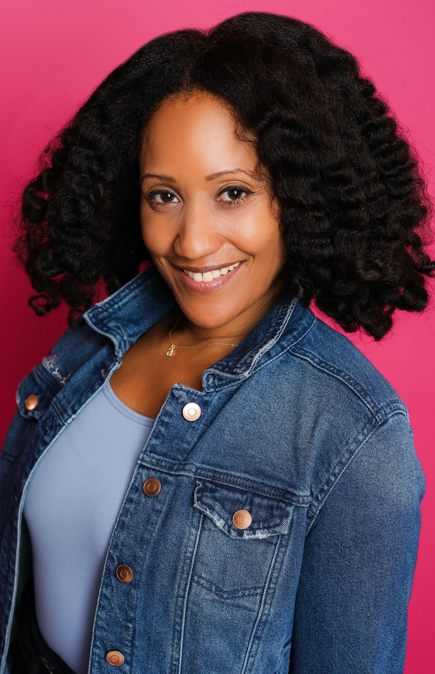 Person in denim jacket with natural curly black hair smiling against pink background.