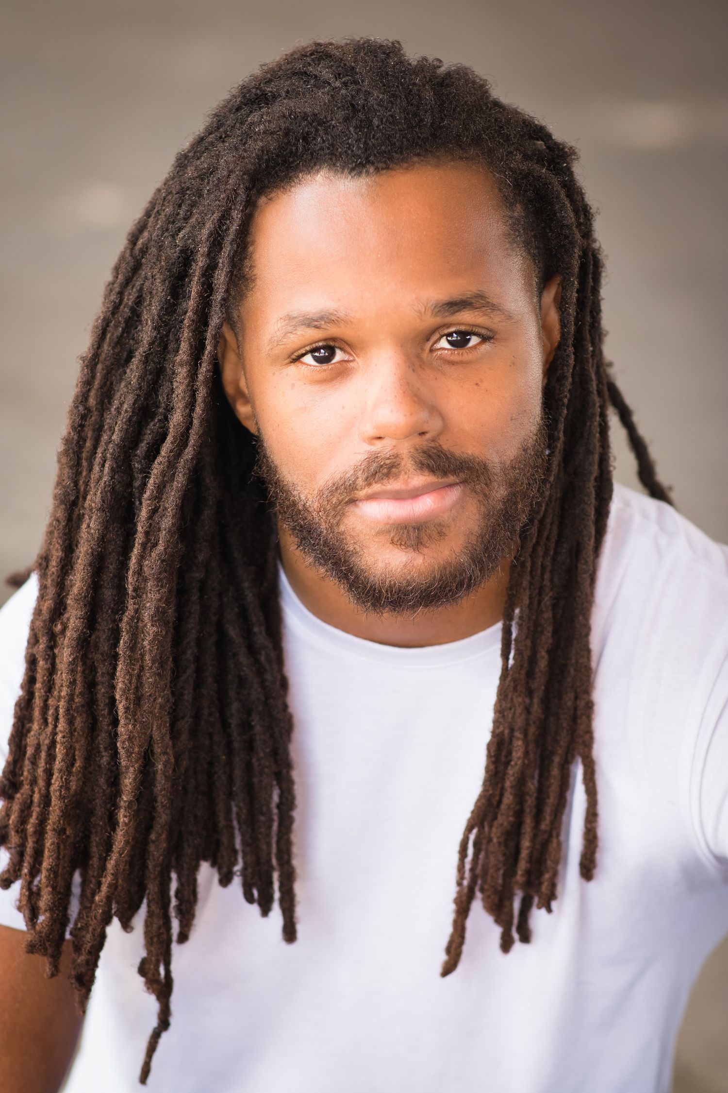 Professional portrait of a person with long dreadlocks wearing a white t-shirt against neutral background.