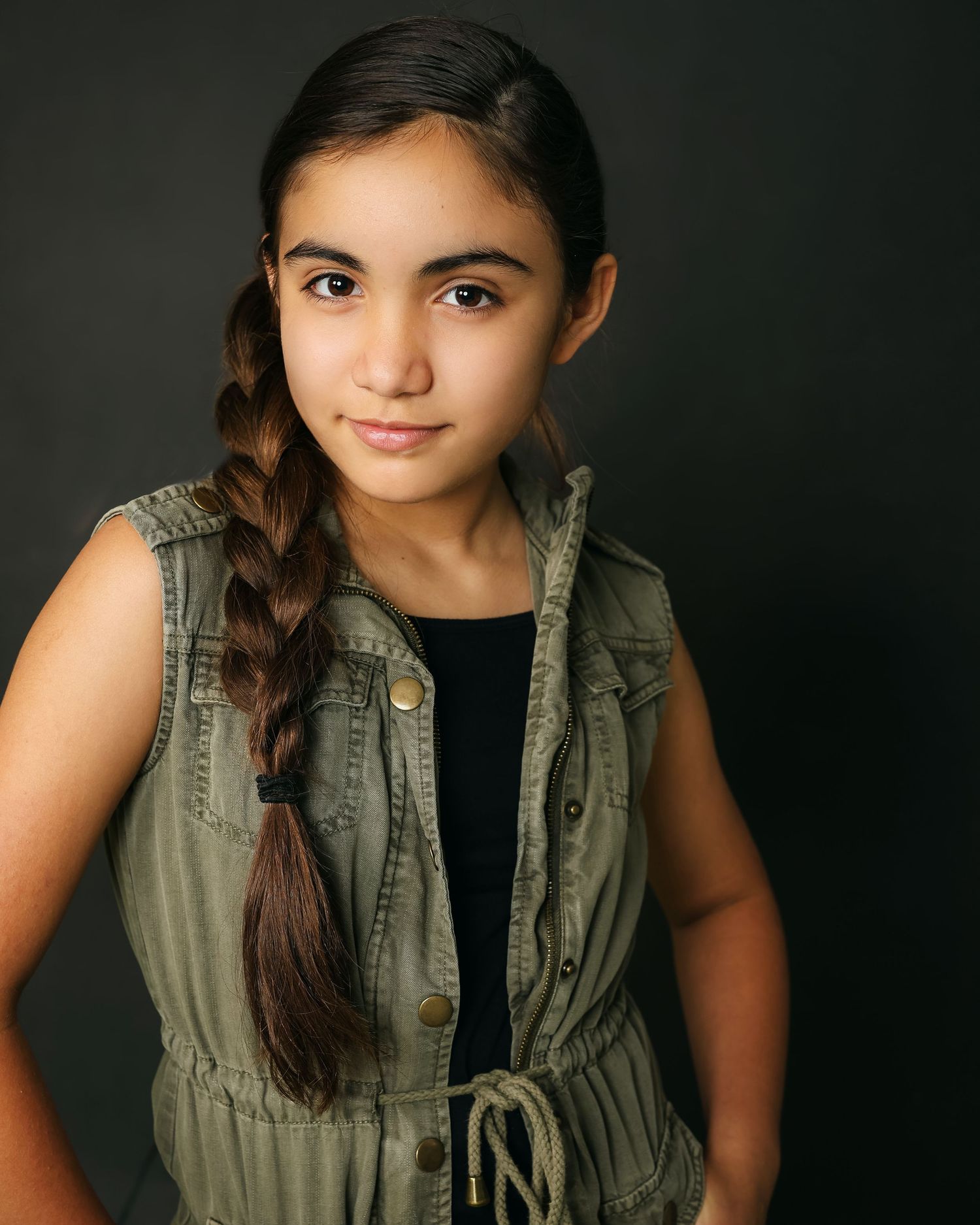 Studio portrait with olive green sleeveless vest over black top and side braid against dark background.