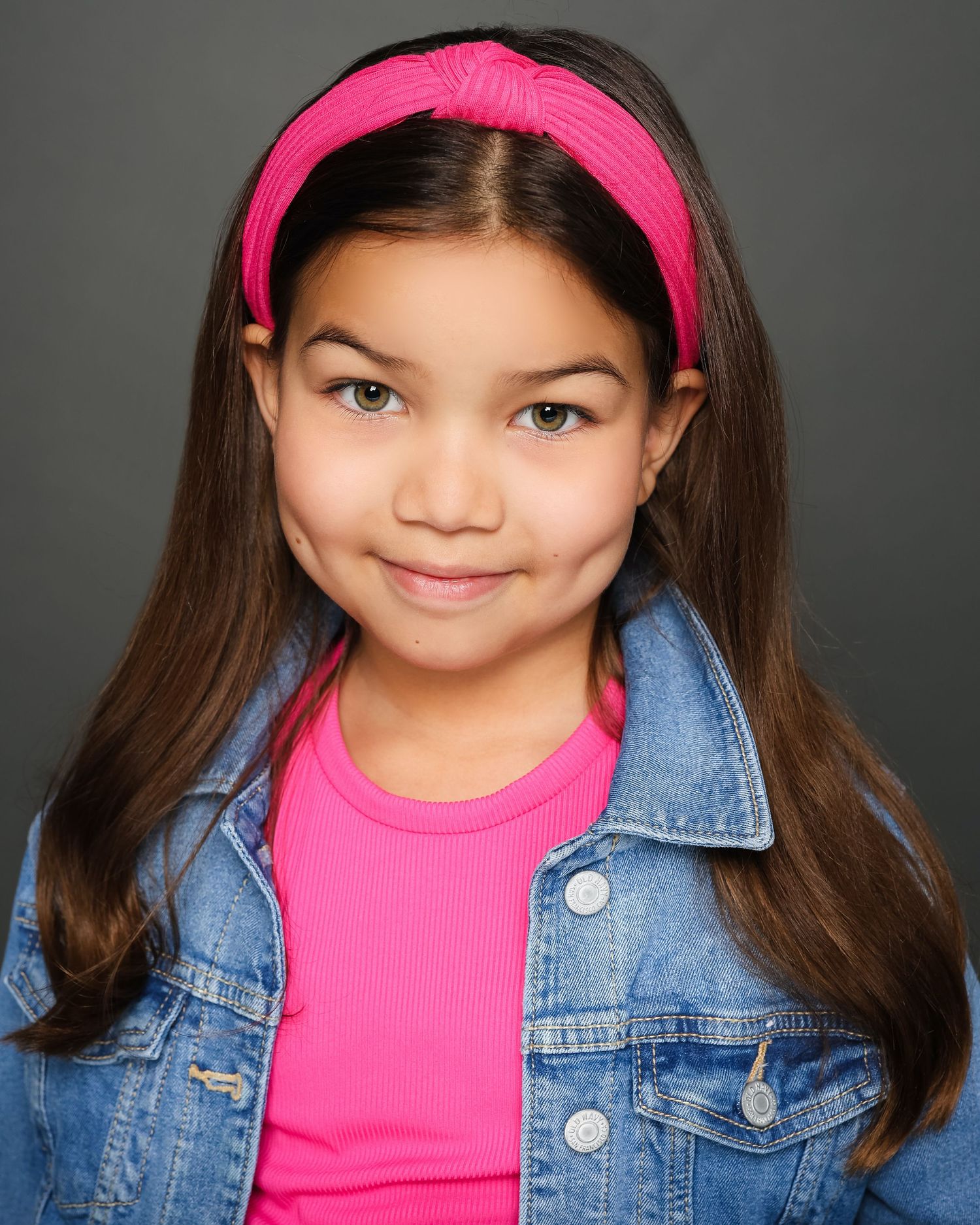 Young model in a denim jacket and bright pink headband poses against a gray backdrop for a professional headshot.