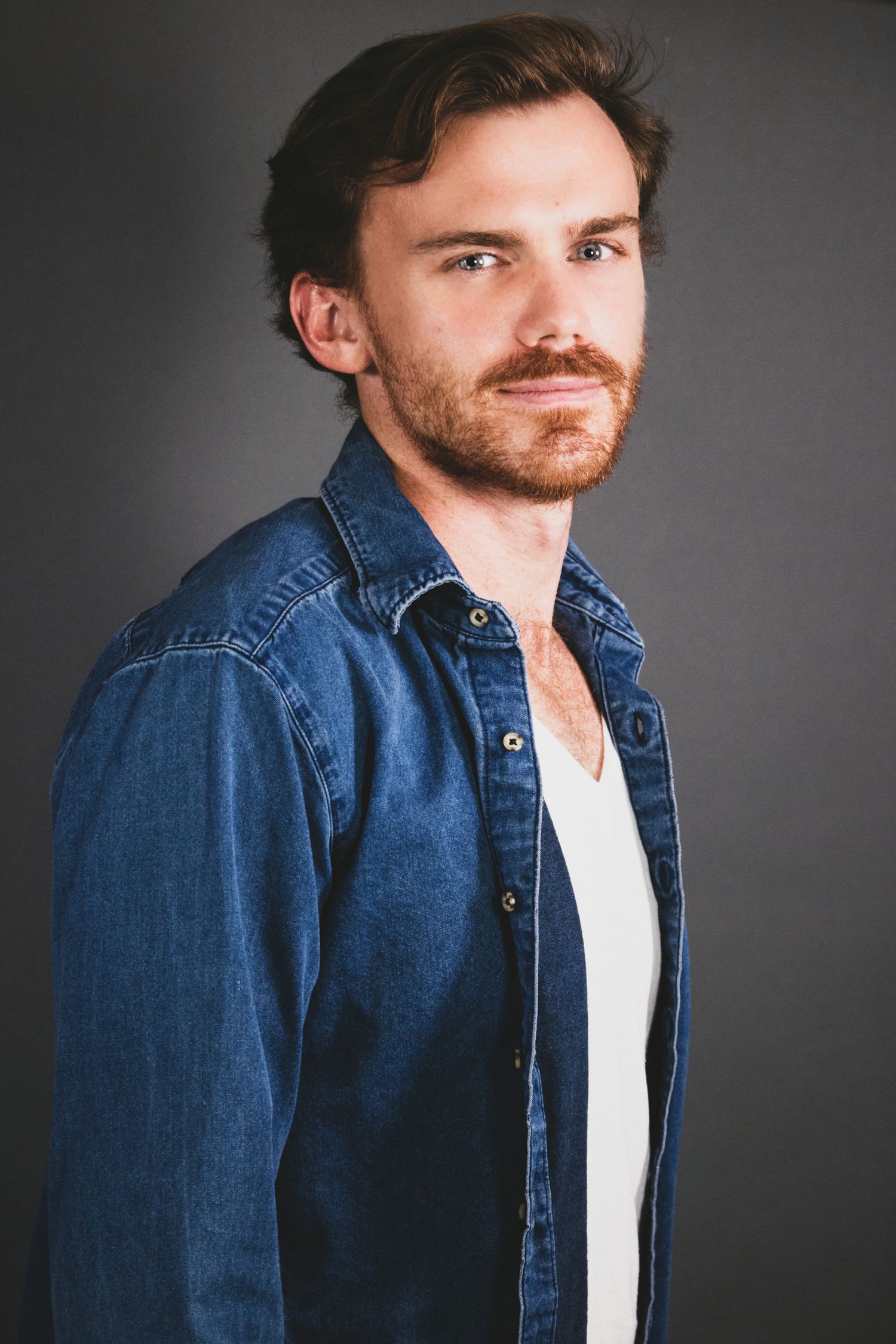 Studio portrait of a young man in denim jacket and white shirt against dark background.