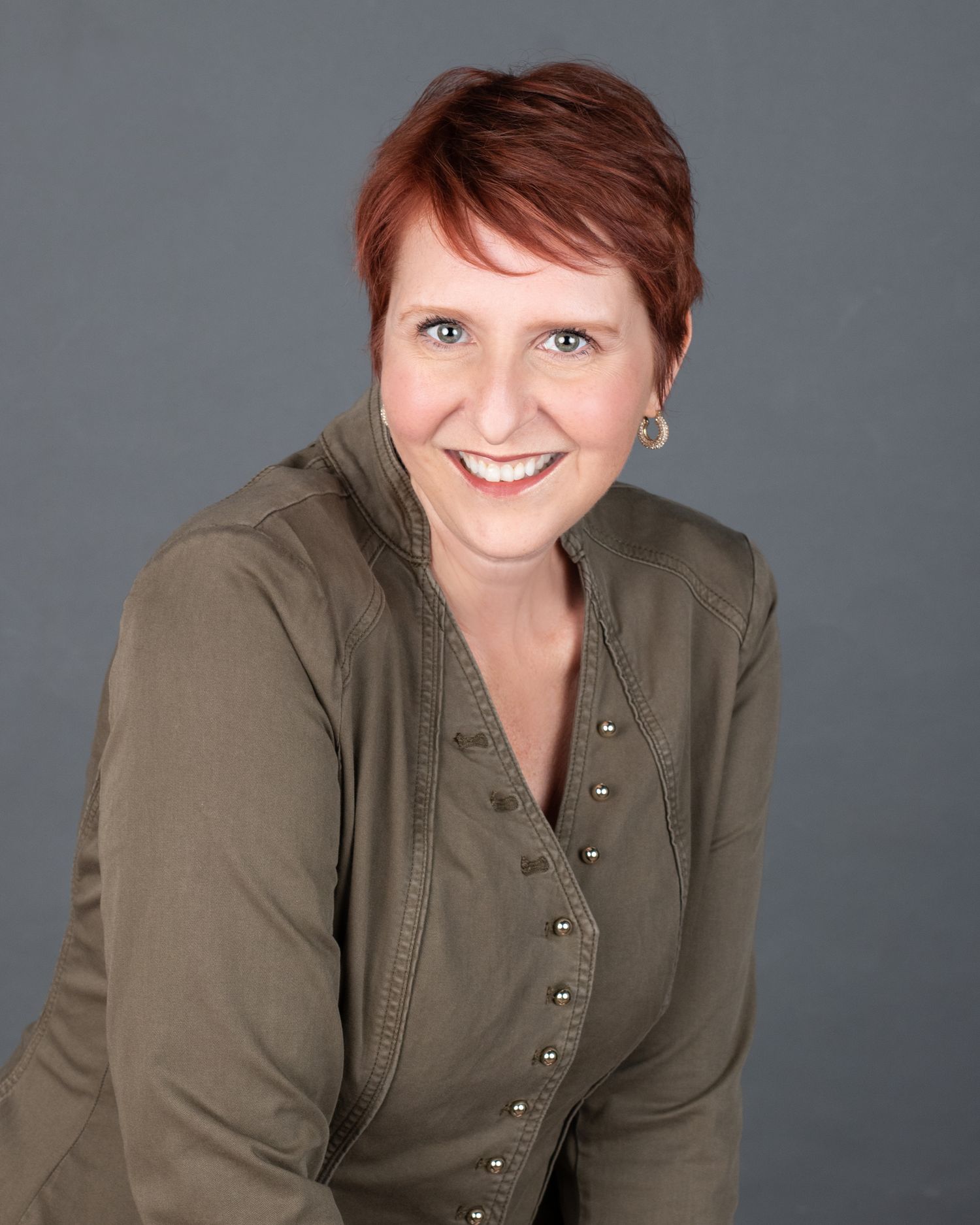 Professional headshot of a person in an olive jacket with short red hair and bright smile.