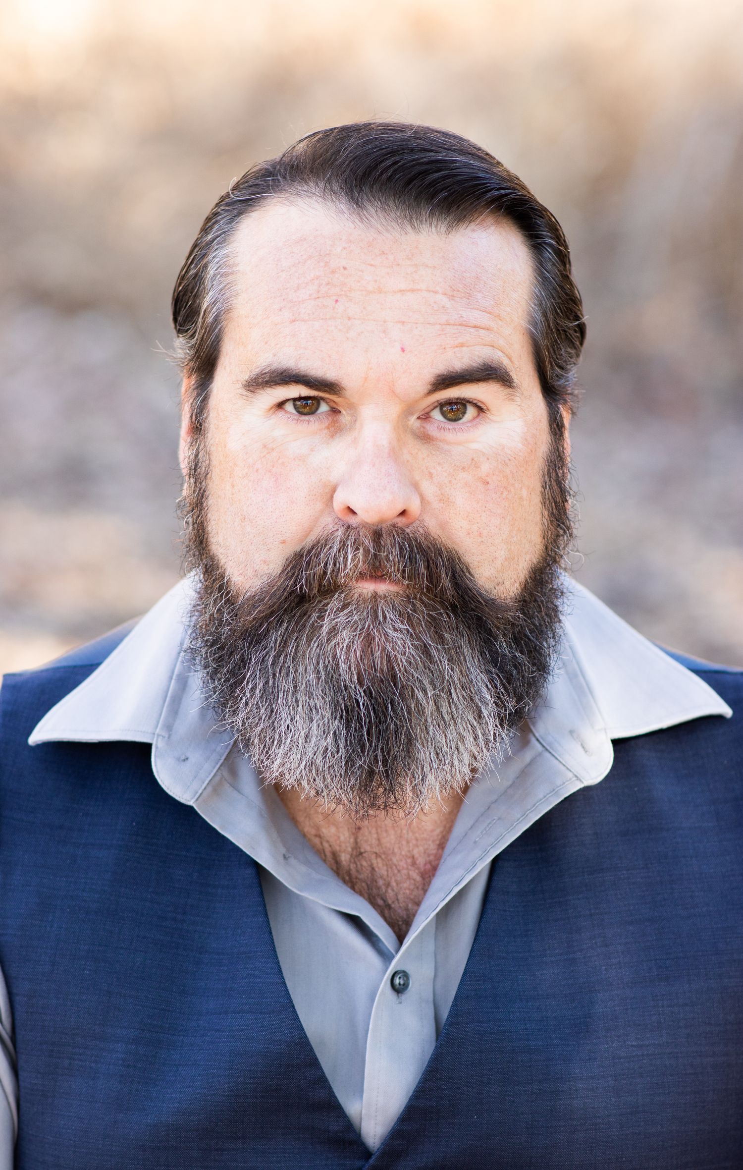Professional headshot of a person with a full beard wearing a navy vest and gray shirt.