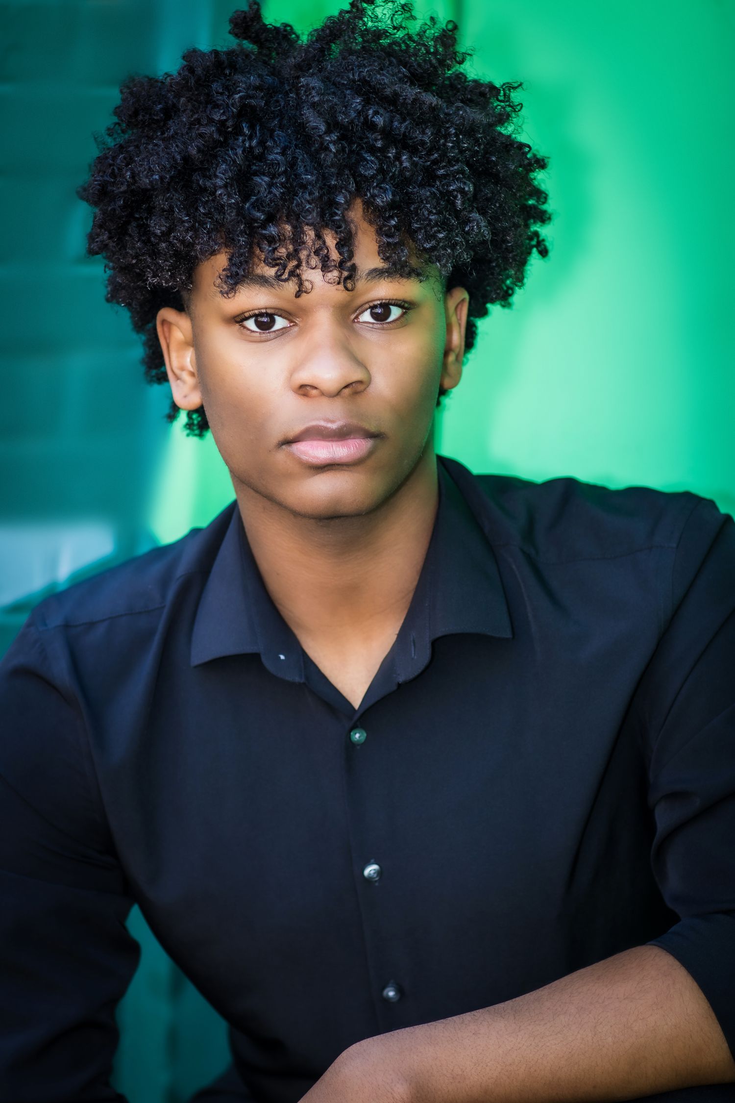 Professional headshot of a person with natural curly hair in dark shirt against teal background.