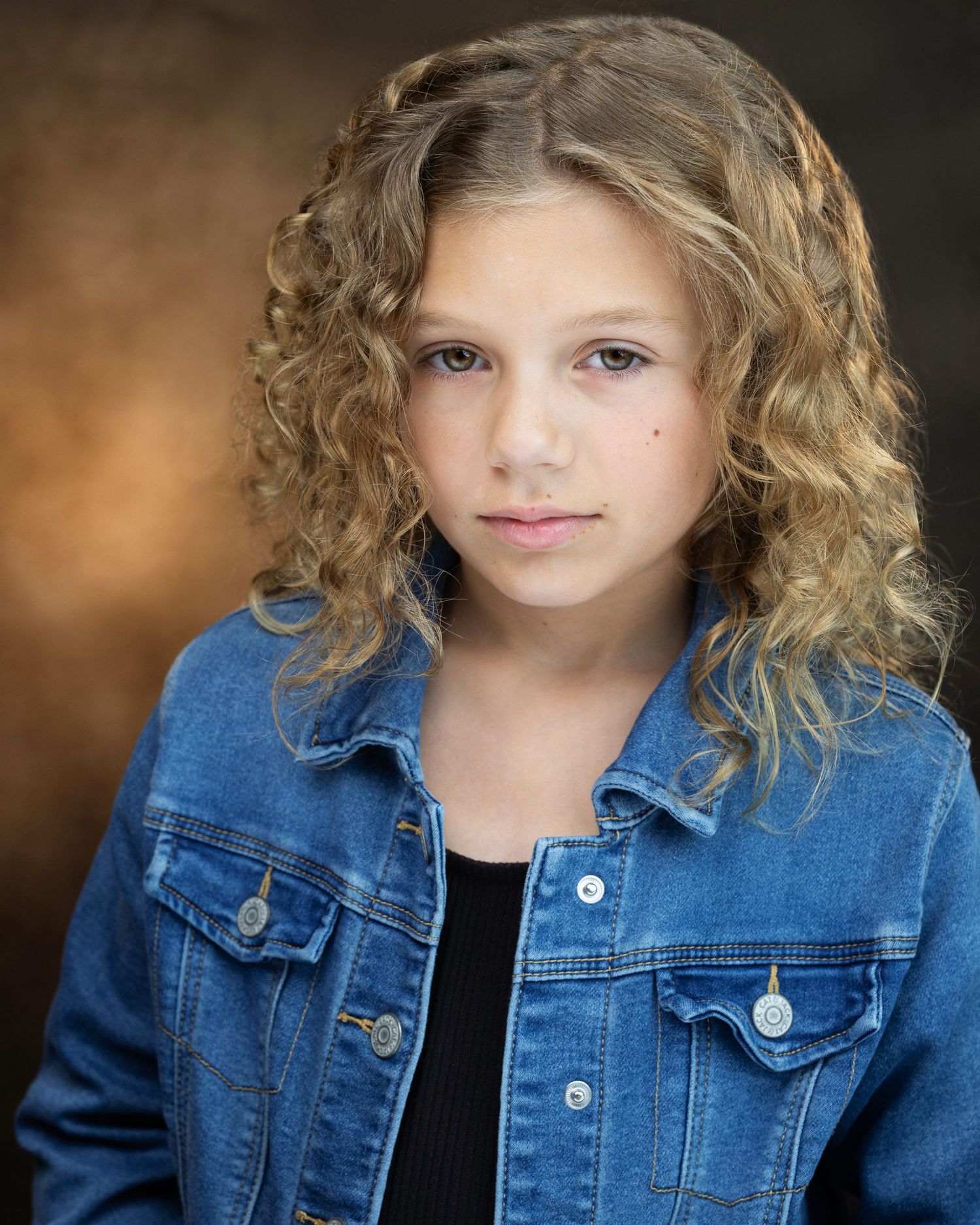 Young girl with curly blonde hair wearing a denim jacket poses for a professional headshot photo.