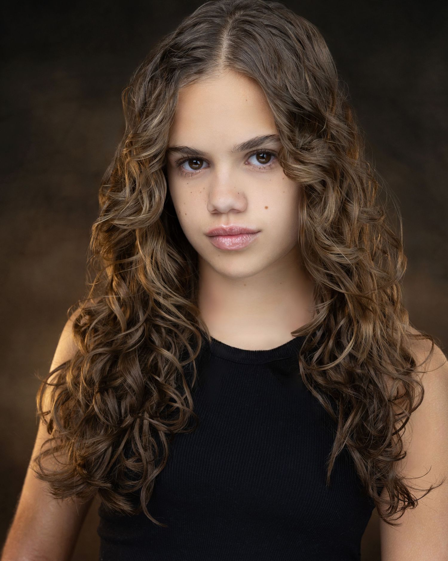Young girl with long curly brown hair wearing a black top, professional headshot portrait photo.