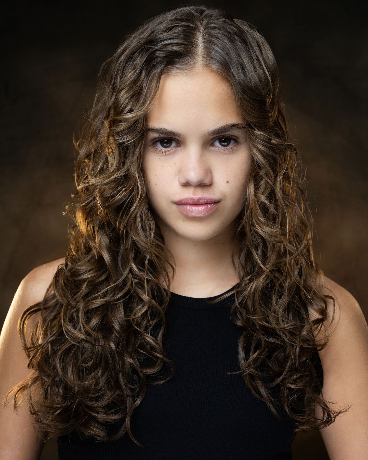 Young girl with long curly brown hair wearing a black top, posing for a professional headshot portrait.