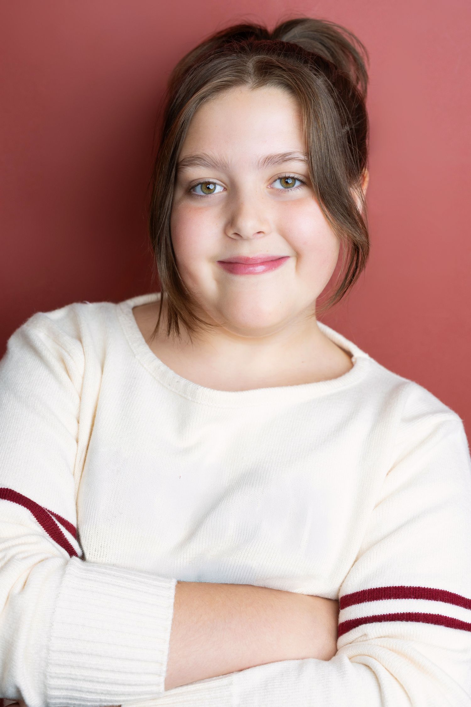 Smiling teenage girl with brown hair in a white sweater with red stripes, arms crossed against a red background.