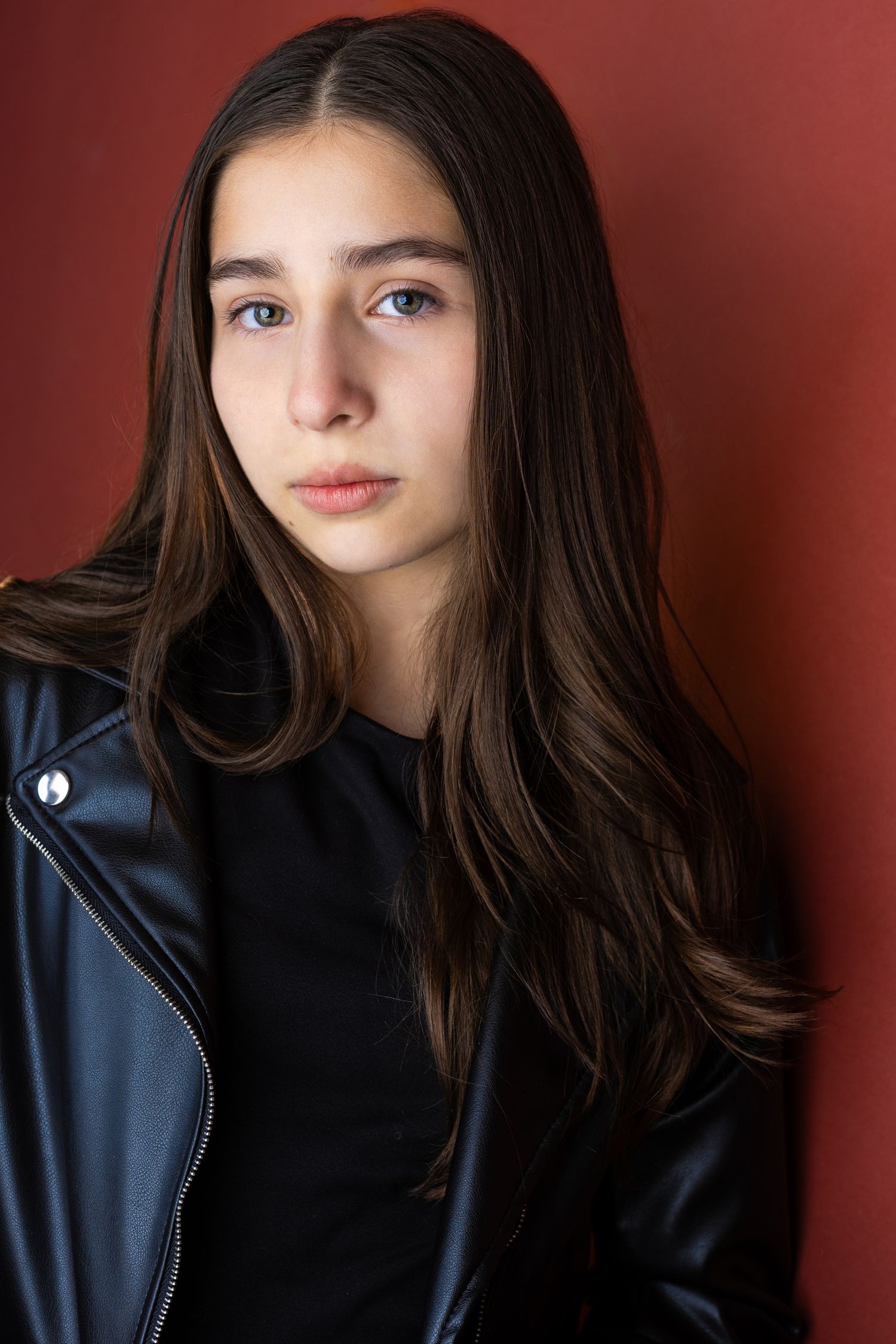 Young teen girl with long dark hair wearing a black leather jacket poses against a red background.