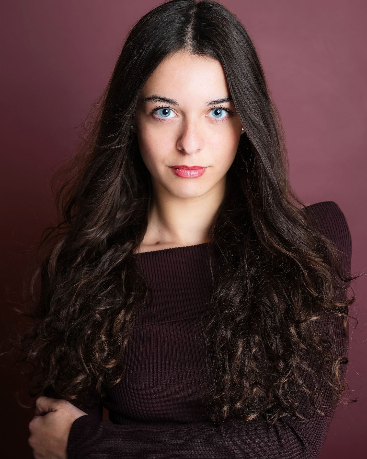 Person in dark sweater with wavy brown hair poses against burgundy backdrop showing different expressions.