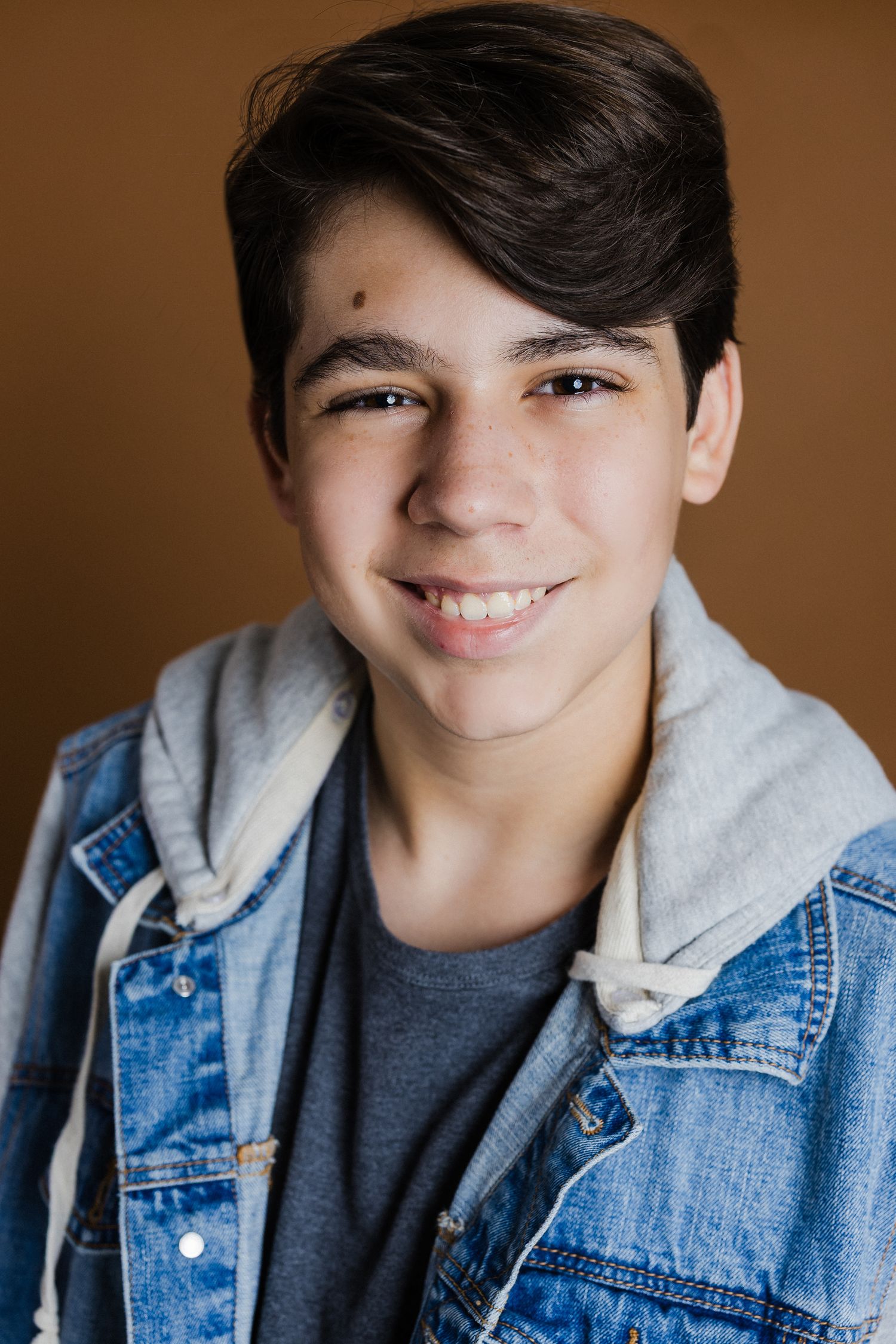 Portrait series in brown studio showing someone wearing denim jacket with grey hood against warm background.