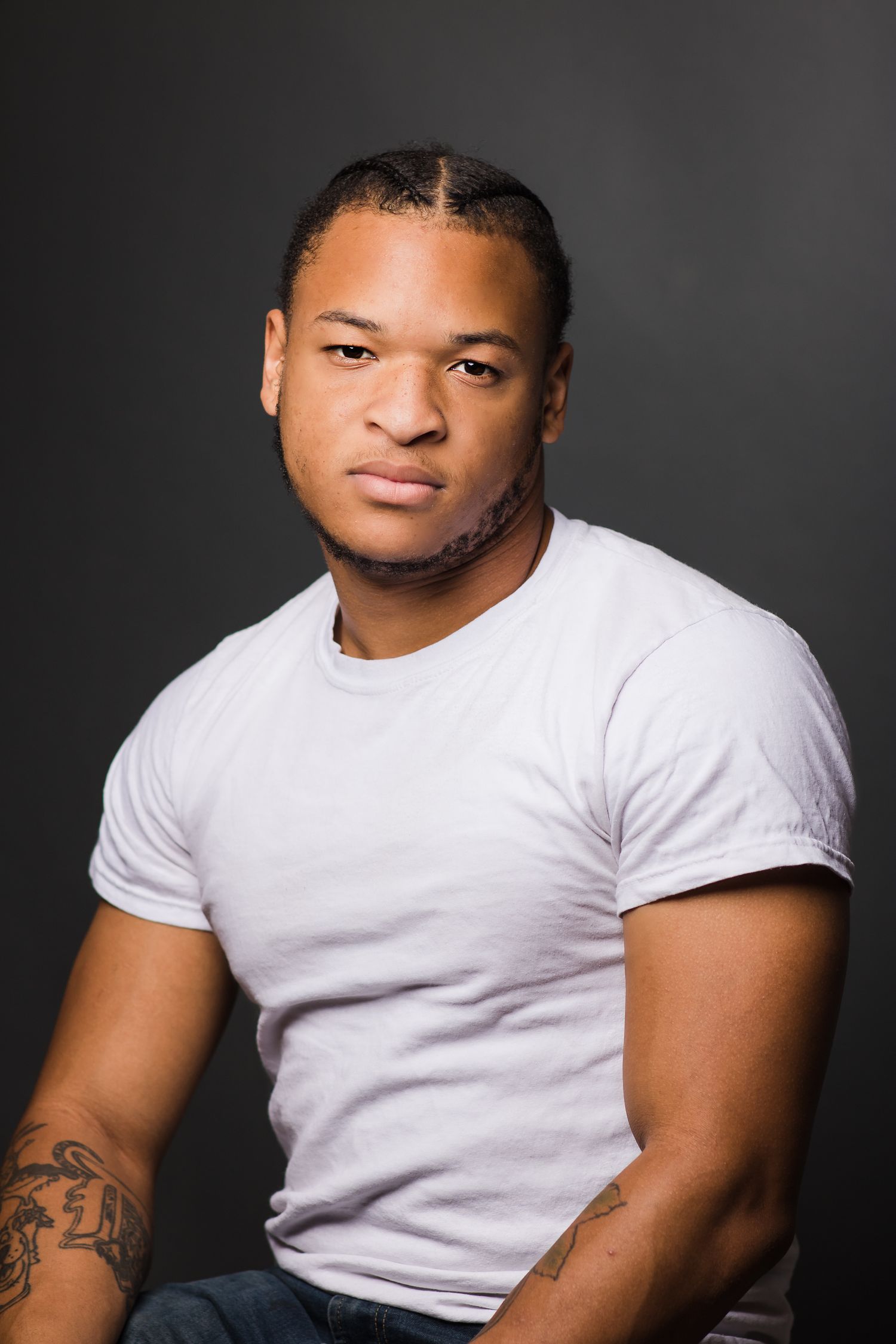 Professional headshot of a muscular person in a white t-shirt against a dark background.