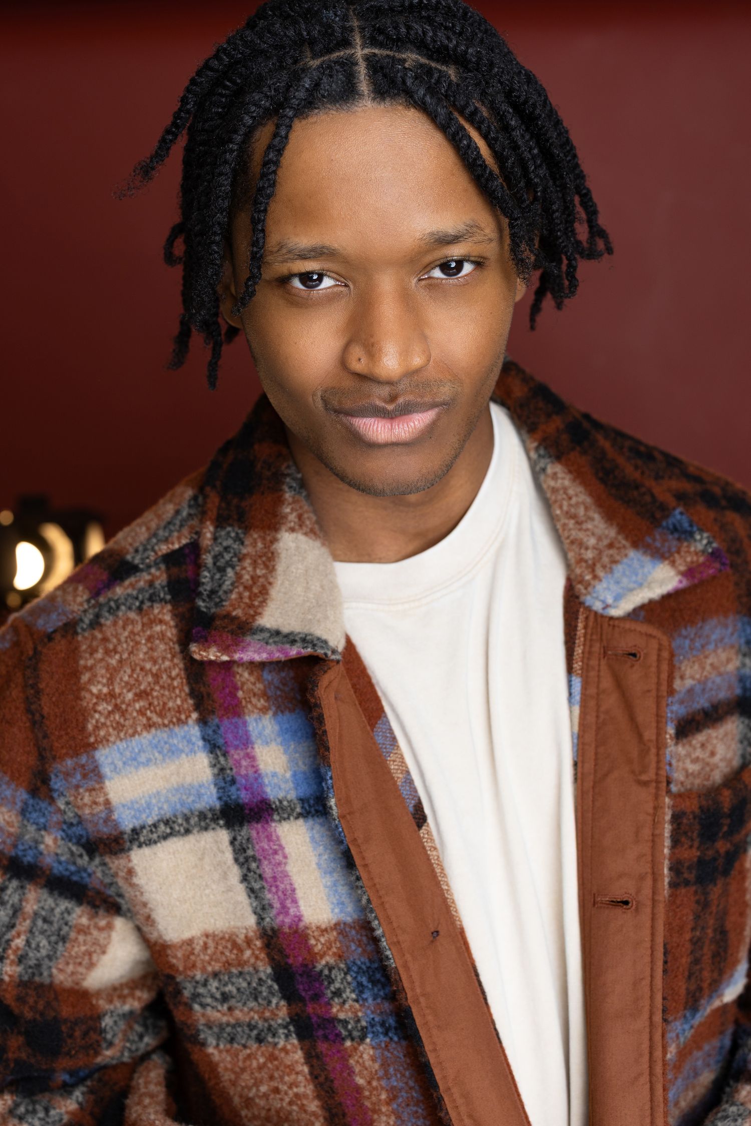 Young Black man with dreadlocks wearing a plaid wool coat over a white shirt, posing against a dark red background.