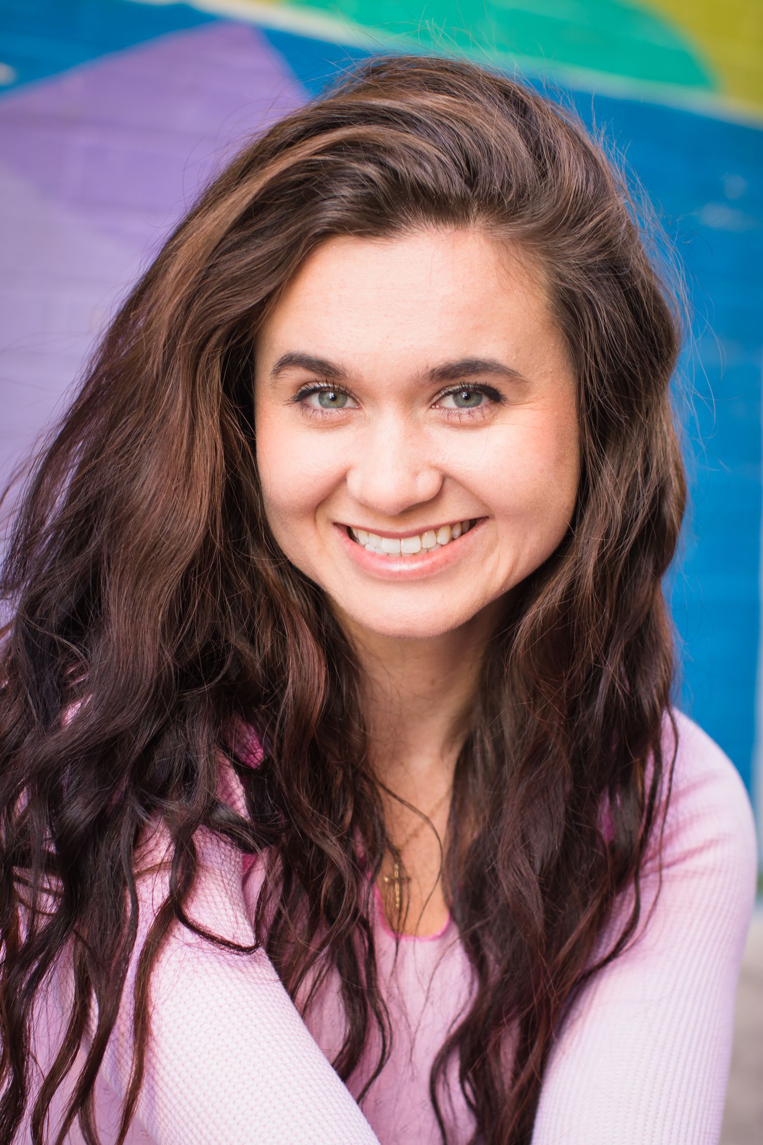 Cheerful portrait of a person with long dark wavy hair wearing pink against a colorful background.