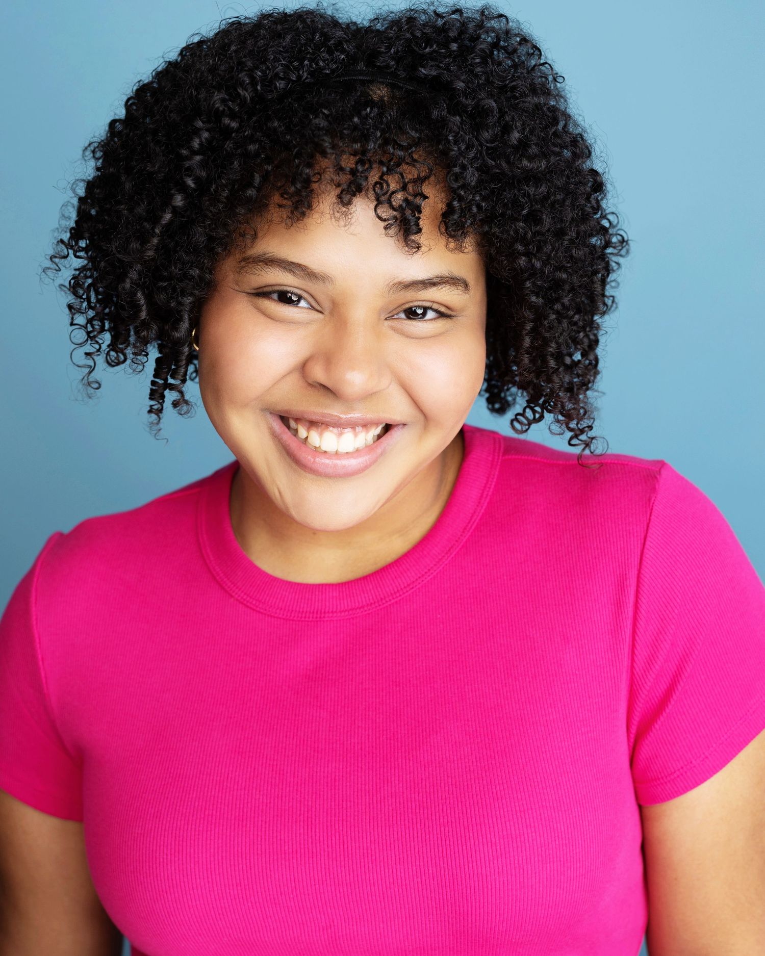Young woman with curly hair smiling brightly, wearing a pink t-shirt against a blue background.