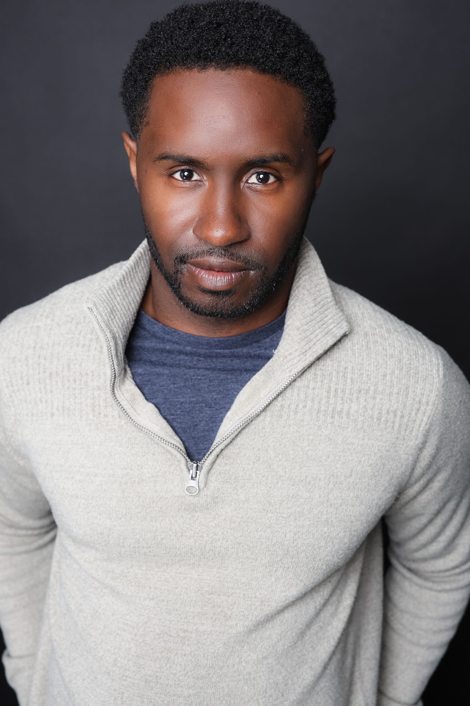 Professional headshot portrait of a person wearing a beige quarter-zip sweater over a navy t-shirt against dark background.