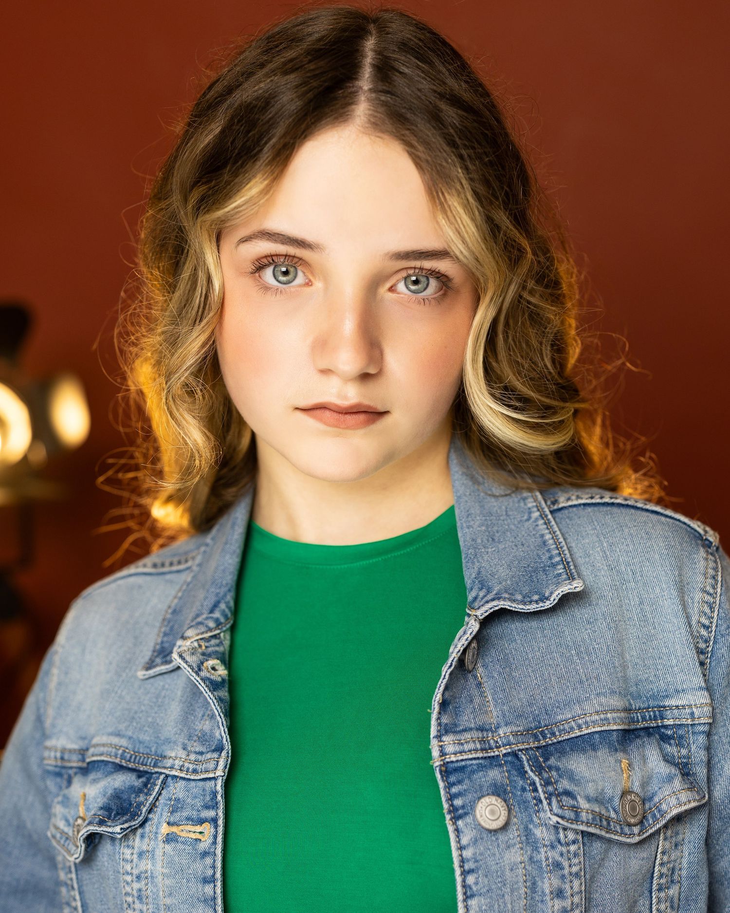 Young girl with blue eyes, wavy hair, green shirt, and denim jacket poses for a professional headshot photo.